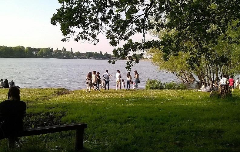 Groups of people of all ages enjoying being in a park. There is a large lake in the background. People are sitting on the grass and standing looking at the water. It is a sunny day.